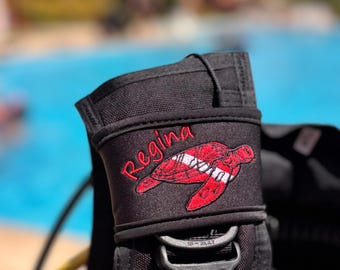 May include: A black scuba diving buoyancy compensator strap featuring an embroidered red sea turtle design with a white stripe across its shell. The name Regina is embroidered in red script above the turtle graphic on the black fabric.