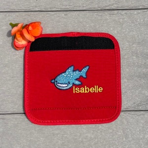 May include: Red fabric square with a black velcro closure. The square has an embroidered whale shark with blue and white details and the name "Isabelle" in yellow thread.