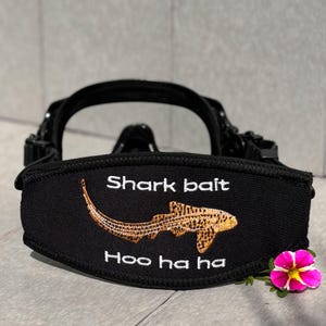 Puede incluir: Una funda de neopreno negro para correa de máscara de buceo con un gráfico bordado de un tiburón leopardo de color marrón dorado. El texto "Shark bait" está bordado en blanco sobre el tiburón y "Hoo ha ha" debajo del gráfico.