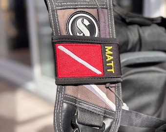 May include: A black scuba diving buoyancy compensator shoulder strap featuring a rectangular red patch with a white diagonal stripe and the name MATT embroidered in yellow thread, positioned below a circular black and white logo emblem.