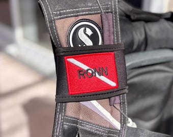 May include: A black scuba diving buoyancy compensator shoulder strap featuring a rectangular red and white diver down flag patch with the name RONN embroidered in black letters, positioned below a circular black and white logo emblem.