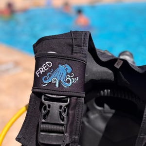Op de afbeelding: Zwarte duikuitrusting met een blauwe octopus patch met de naam "FRED". De octopus is geborduurd in blauwe en turquoise tinten. Een zwarte gesp en gele slang zijn ook zichtbaar.