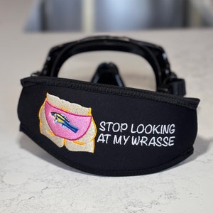 Puede incluir: Máscara de buceo negra con una funda de correa de neopreno negra. La funda presenta un diseño bordado de una forma rosa con un pez azul y amarillo, y el texto "STOP LOOKING AT MY WRASSE" en blanco.
