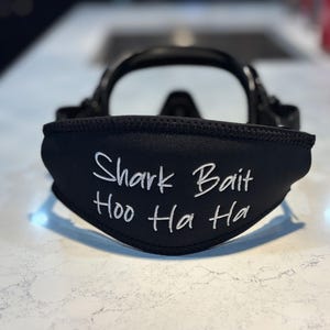 Könnte beinhalten: Eine schwarze Tauchmasken-Bandabdeckung aus Neopren mit dem weißen, gestickten Text "Shark Bait Hoo Ha Ha" in der Mitte. Die Abdeckung ist an einem schwarzen Tauchmaskenrahmen befestigt, wobei ein klares Glas hinter der Abdeckung sichtbar ist.