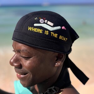 A black bandana with the text "WHERE IS THE BOAT" embroidered in yellow. The bandana features a small embroidered design of a person swimming with a sad face and a life preserver.