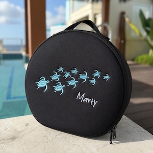 May include: A round black carrying case featuring an embroidered pattern of light blue sea turtles swimming across the front. The name Marty is embroidered in white script lettering below the sea turtle design. The case has a top handle and zipper.