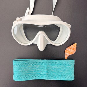 May include: White diving mask with a clear lens and a blue headband. The mask has the word "TEMPERED CE" printed on it.