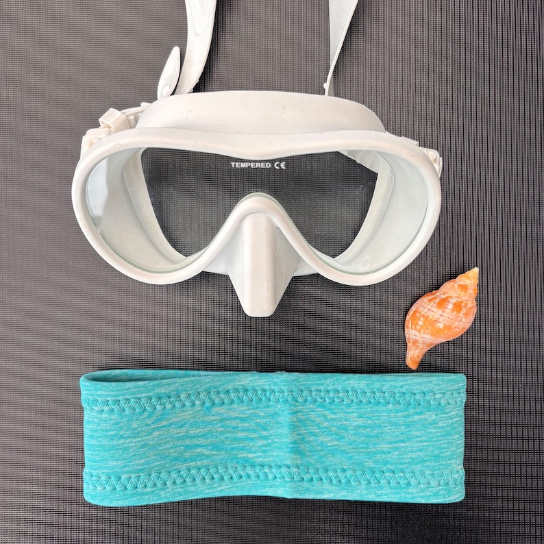 May include: White diving mask with a clear lens and a blue headband. The mask has the word "TEMPERED CE" printed on it.