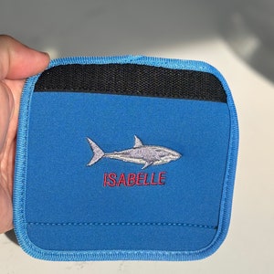 Puede incluir: Una cartera azul con un diseño de tiburón bordado en gris y blanco. El nombre "ISABELLE" está bordado en rojo debajo del tiburón. La cartera tiene una parte superior negra y ribetes azules.