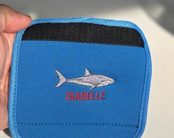 May include: A square blue neoprene pouch featuring a black fabric strip at the top edge. The center displays an embroidered grey shark graphic positioned above the name ISABELLE stitched in red thread. The edges are finished with blue piping.
