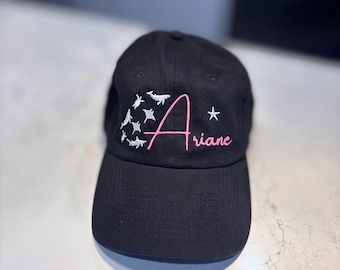 May include: Black baseball cap with a white star and a pink cursive name "Ariane" embroidered on the front. The cap also features a white embroidered design of a whale and other sea creatures.