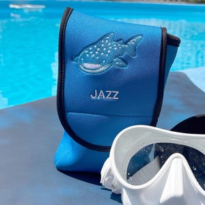 May include: A blue scuba mask case with a black trim and the word "JAZZ" printed on it. The case features an embroidered whale shark design. A white scuba mask is partially visible in the foreground.