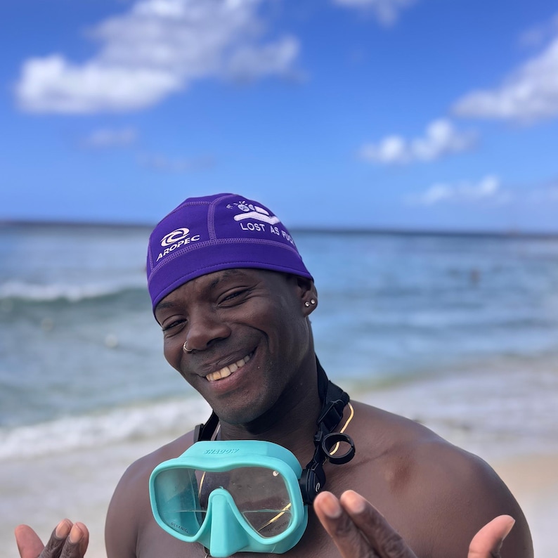 A man wearing a purple swim cap with the text "LOST AS F*#K" and a turquoise diving mask. The man is smiling and has a gold chain around his neck.