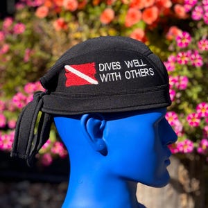 May include: Black scuba diving cap with the text "DIVES WELL WITH OTHERS" in white. The cap features a red and white scuba flag on the left side. The cap is tied in the back and is displayed on a blue mannequin head.