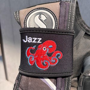 May include: Black fabric accessory with the name "Jazz" in white script and a red octopus with blue accents. The accessory is attached to a larger, grey and black item with a silver emblem.