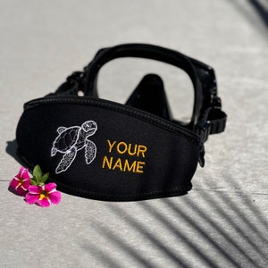 Op de afbeelding: Een zwarte neopreen duikmaskerbandhoes met een geborduurde witte zeeschildpad en de gele tekst YOUR NAME. De bandhoes is bevestigd aan een zwart duikmasker, met twee kleine roze en gele bloemen ernaast geplaatst op de ondergrond.