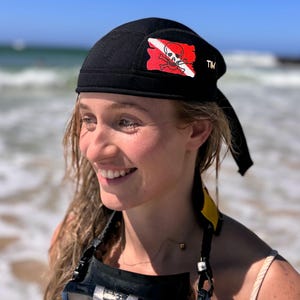 May include: Black scuba diving cap with a red and white flag design featuring a skull and crossbones. The cap has a curved brim and a strap that goes under the chin. The cap is worn by a person on a beach.