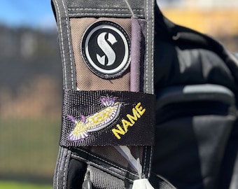 May include: A black scuba buoyancy compensator strap featuring a circular logo with a white letter S. A black fabric patch is attached, displaying an embroidered yellow and purple nudibranch sea slug graphic and the word NAME in yellow.