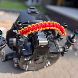 Könnte beinhalten: Schwarzes Unterwasserkameragehäuse mit einem rot-orangenen geflochtenen Griff. Die Kamera hat mehrere Knöpfe und Tasten. Der Griff ist mit Metallbeschlägen an der Oberseite des Gehäuses befestigt.