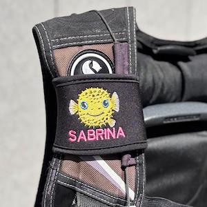 Pode incluir: Mochila preta com uma alça listrada preta e branca. Uma bolsa preta com um baiacu amarelo e o nome "SABRINA" bordado em rosa está presa à alça.