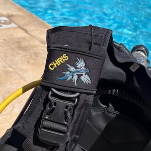 May include: A black scuba diving buoyancy compensator vest featuring a yellow embroidered name tag that reads CHRIS and an embroidered blue and white sea slug graphic on the shoulder strap, complete with a black plastic buckle.