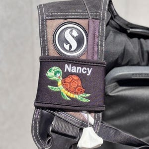May include: A black scuba diving gear bag with a brown strap and a black patch with a white letter S. The bag has a black sleeve with a green and brown turtle graphic and the name "Nancy" embroidered in white.