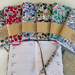 Op de afbeelding: Een collectie van vijf met stof beklede planners met bloemmotieven in rood, blauw, groen en paars, elk omwikkeld met een bruine papieren band met het label 100% FAIT MAIN ORIGINAL. Een open 2025-planner ligt eronder met een bijpassend potlood.