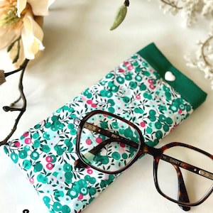 May include: A floral print glasses case with a green border and a white heart-shaped button. Tortoiseshell eyeglasses rest on the case. The case features a pattern of green and pink flowers on a white background.