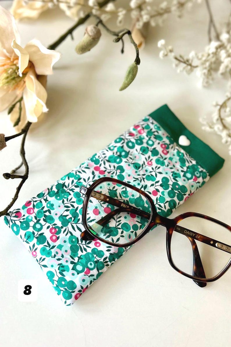 May include: A floral print glasses case with a green border and a white heart-shaped button. Tortoiseshell eyeglasses rest on the case. The case features a pattern of green and pink flowers on a white background.