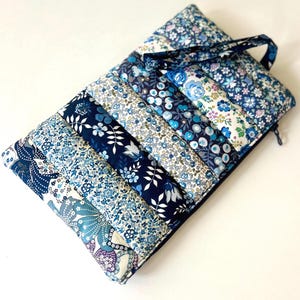 Puede incluir: Un estuche de tela acolchada en varios tonos de azul, con estampados florales. El estuche tiene un asa y un cierre de cremallera. El diseño incluye una variedad de estampados florales azules, creando un efecto patchwork. El estuche mide unos 25 cm de largo.