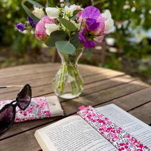 May include: A vibrant floral arrangement in a glass vase on a wooden table. A book with a floral bookmark, sunglasses, and a matching glasses case are also present. The flowers are pink, purple, and white.
