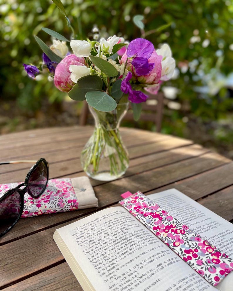 May include: A vibrant floral arrangement in a glass vase on a wooden table. A book with a floral bookmark, sunglasses, and a matching glasses case are also present. The flowers are pink, purple, and white.