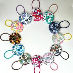 Peut inclure: Un ensemble de 15 élastiques à cheveux colorés avec des motifs floraux. Chaque élastique est doté d'un bouton rond avec un motif floral différent. Les boutons sont en tissu et ont un fond blanc avec des fleurs et des feuilles de différentes couleurs.