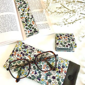 May include: An eyeglasses case, bookmark, and small notebook are shown. The case and bookmark have a floral pattern with green, blue, and purple flowers on a white background. The eyeglasses have a brown tortoiseshell frame.