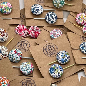 Könnte beinhalten: Eine Sammlung handgefertigter Haarklammern mit verschiedenen floralen Stoffknöpfen. Jede Klammer ist an einem Holzstab befestigt und hat ein braunes Papieretikett mit dem aufgedruckten Text "100% FAIT main ORIGINAL".