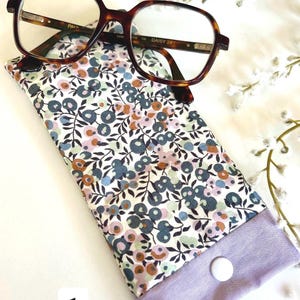 May include: Tortoiseshell-framed spectacles sit atop a floral glasses case. The case displays a pattern of blue, pink, and brown flowers on a white backdrop, with a lavender closure fastened by a white button.