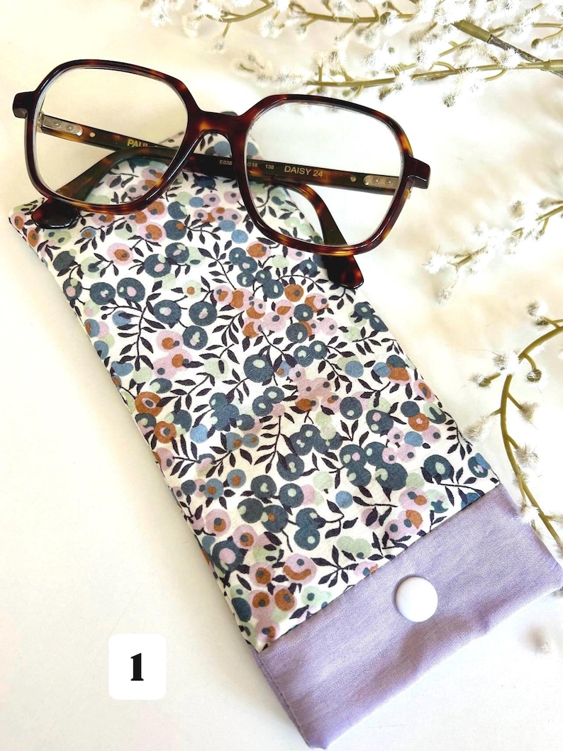May include: Tortoiseshell-framed spectacles sit atop a floral glasses case. The case displays a pattern of blue, pink, and brown flowers on a white backdrop, with a lavender closure fastened by a white button.