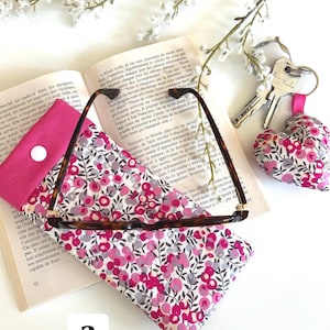 May include: A glasses case, a small coin purse, and a heart-shaped keychain are displayed on an open book. The glasses case and keychain are made of a floral fabric with pink and grey flowers. The glasses are tortoise shell.