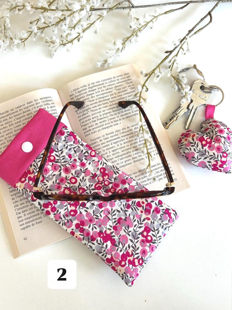 May include: A glasses case, a small coin purse, and a heart-shaped keychain are displayed on an open book. The glasses case and keychain are made of a floral fabric with pink and grey flowers. The glasses are tortoise shell.
