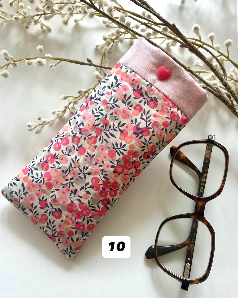 May include: A floral eyeglasses case with a pink top and a heart-shaped button. The case is next to a pair of brown, square-framed eyeglasses. The case has a pattern of pink and red flowers on a cream background. The number "10" is in the lower left corner.