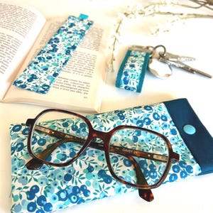 May include: A book with a blue floral bookmark, glasses, and a matching glasses case. A key fob with a similar floral pattern is also present. The glasses have a tortoiseshell frame. The number "3" is visible.