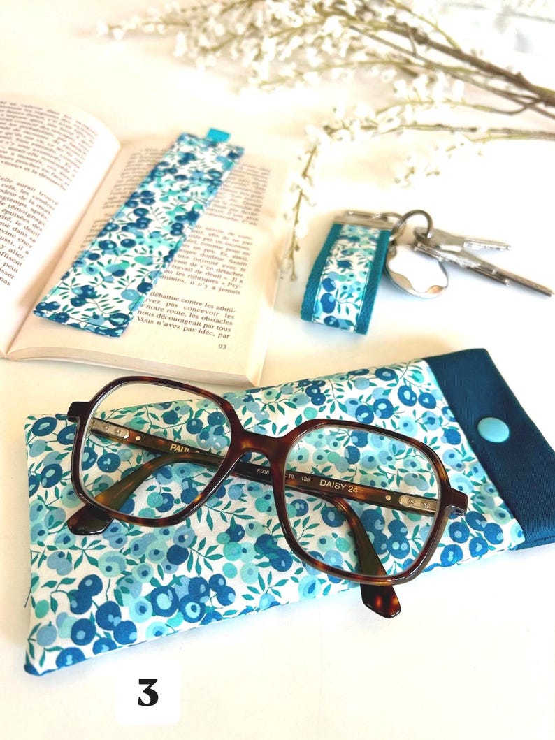 May include: A book with a blue floral bookmark, glasses, and a matching glasses case. A key fob with a similar floral pattern is also present. The glasses have a tortoiseshell frame. The number "3" is visible.