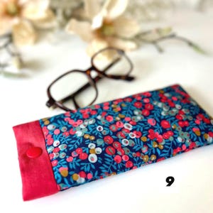 May include: A patterned glasses case with a red trim and button. The case has a navy blue floral design with red, pink, and white flowers and green leaves. A pair of tortoiseshell glasses sits nearby on a white surface.