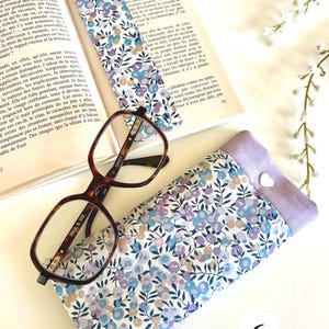 May include: A floral print glasses case, a matching bookmark, and eyeglasses rest on an open book. The case and bookmark feature a pattern of blue, purple, and white flowers. The eyeglasses have a brown tortoiseshell frame.