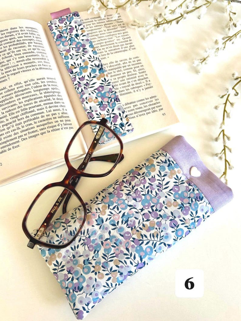May include: A floral print glasses case, a matching bookmark, and eyeglasses rest on an open book. The case and bookmark feature a pattern of blue, purple, and white flowers. The eyeglasses have a brown tortoiseshell frame.