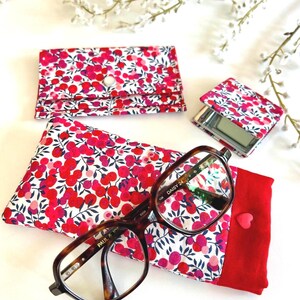May include: A collection of accessories with a floral design in red and pink on white. The items include a glasses case, a compact mirror, and a card holder. A pair of spectacles rests on a matching case.