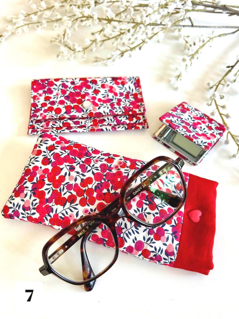 May include: A collection of accessories with a floral design in red and pink on white. The items include a glasses case, a compact mirror, and a card holder. A pair of spectacles rests on a matching case.
