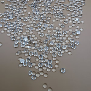 May include: A large quantity of clear glass beads scattered on a light brown surface. The beads are round and have a slight shimmer.