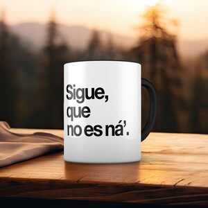 May include: White ceramic mug with a black handle, featuring the Spanish text "Sigue, que no es ná." The mug is set on a wooden surface with a blurred background of trees and sunlight.