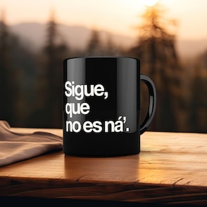 May include: Black ceramic mug with the text "Sigue, que no es ná'" in white. The mug has a curved handle and is sitting on a wooden surface. The background is blurred, showing a natural outdoor scene.